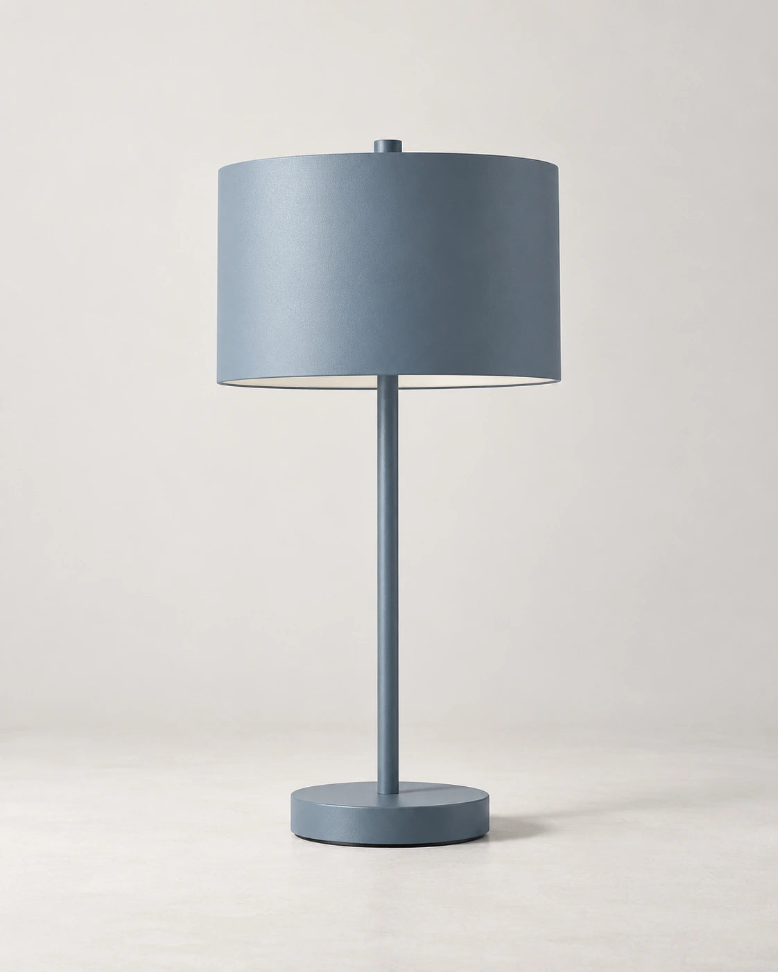 Blue-grey metal table lamp with a slim stem, cylindrical shade and circular base on a light background.