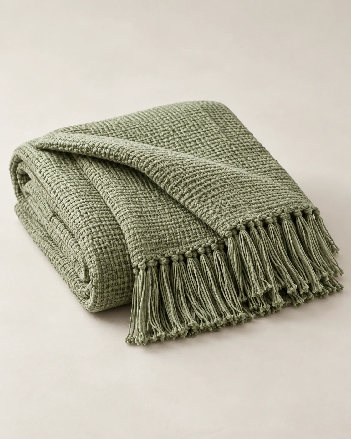Folded sage woven throw with a fringed edge and visible textile texture on a pale background.